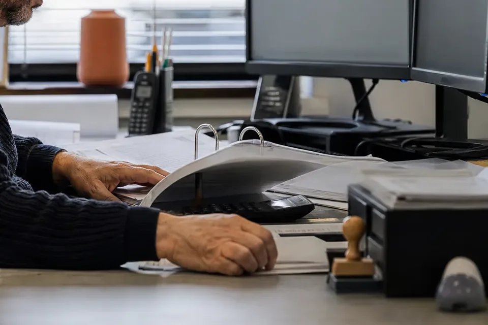 Hands turning pages of a binder of monthly financials next to a calculator at a finance desk