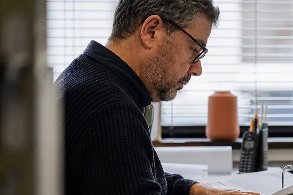 Controller in glasses reading through a binder of reconciliations at an office desk by the window