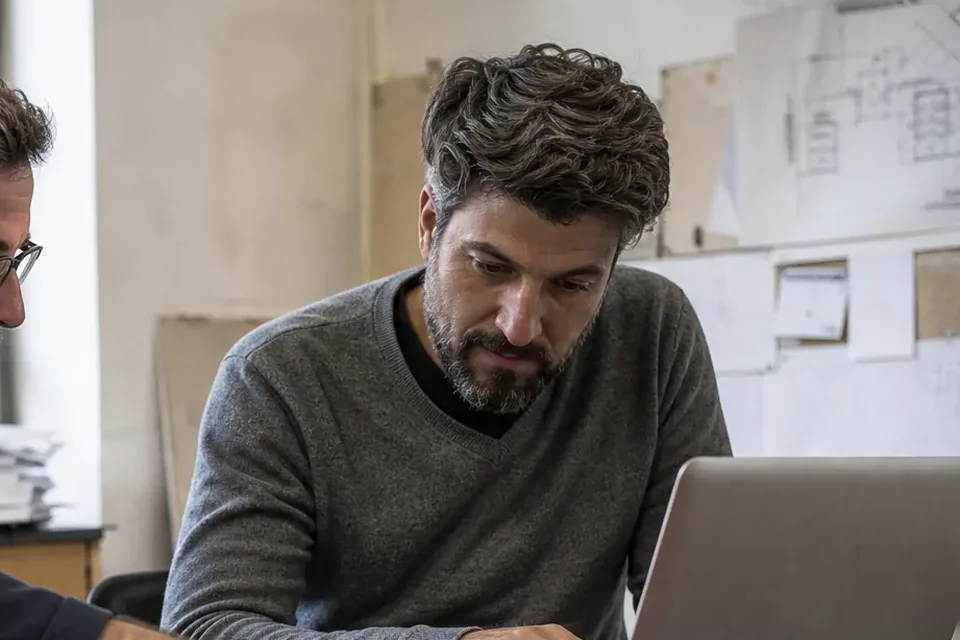 Engineering leader reviewing work on a laptop with architecture sketches pinned on the wall behind him